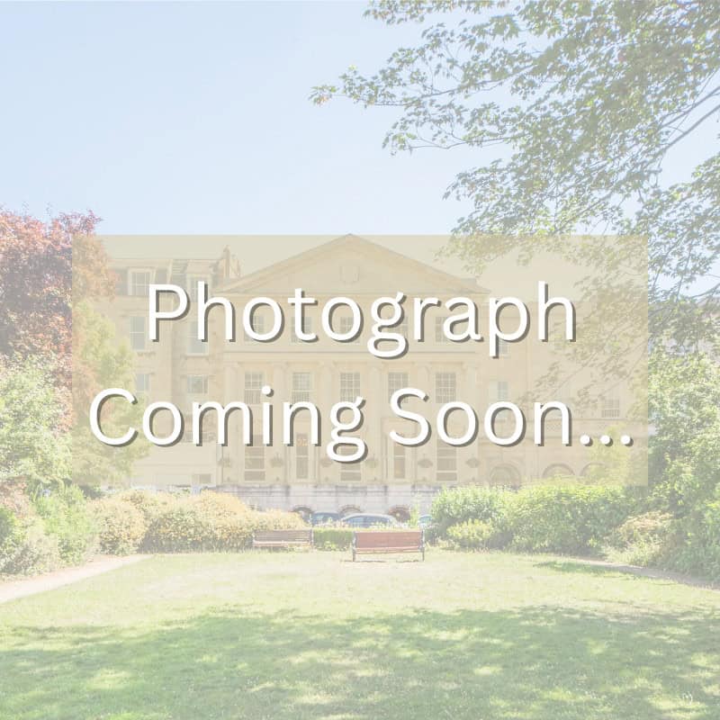 Image placeholder with the text "Photograph Coming Soon" over a blurred outdoor scene featuring a building, trees, and a grassy area with a bench.