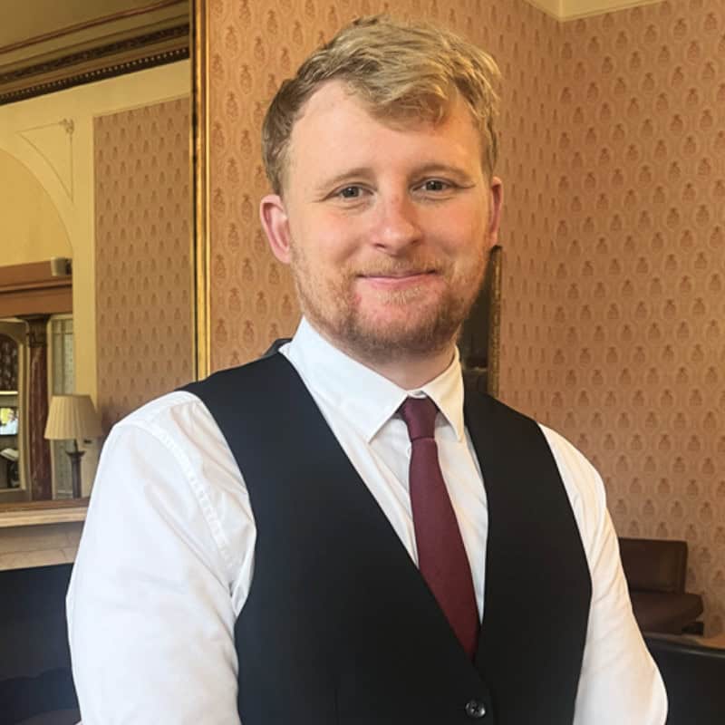 A man with short blond hair wearing a black vest, white shirt, and red tie smiles in a room with patterned wallpaper and a large mirror.