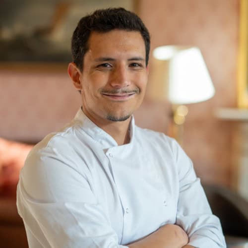 A smiling person wearing a white chef's coat stands with arms crossed in a warmly lit room.