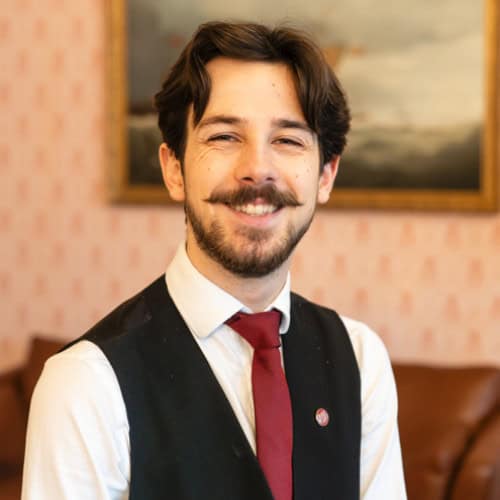 A smiling man with a mustache wearing a white shirt, black vest, and red tie stands indoors, with a painting in the blurred background.