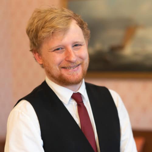 Smiling man with red hair and beard wearing a vest and tie in a warmly lit room with a painting in the background.