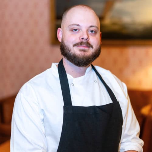 A man with a beard wearing a white chef's jacket and black apron stands indoors, smiling slightly. The background is softly lit.